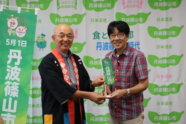 市長が黒い法被を着て、原田さんに丹波篠山市と書かれた小さい旗を渡している写真