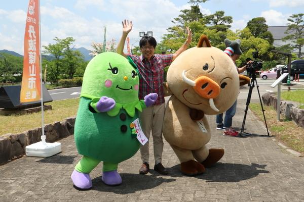 まめりんとまるいのの間で、両手を挙げてポーズをとっている原田さんの写真