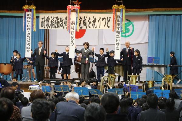 生徒が演奏する前で、舞台上のくず玉の下で手を取り合う生徒と教師と市長の写真