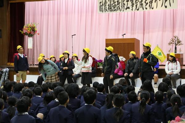 生徒たちの前で、複数の教師がランドセルを背負い、黄色い帽子をかぶって、小学生を演じる写真