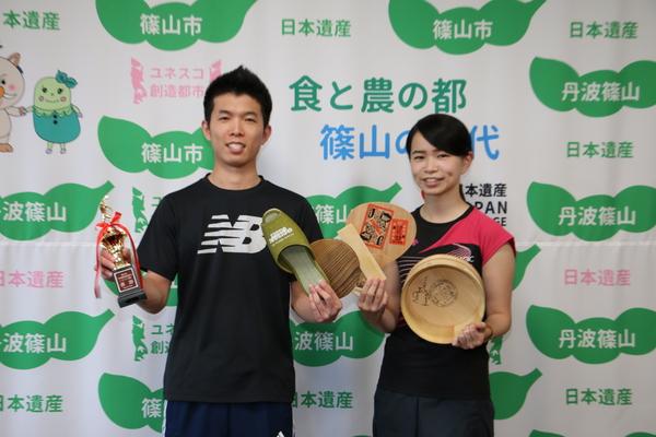 ユニホーム姿の男女ペアの選手が優勝のトロフィーやスリッパ・ラケット・風呂桶などを持って記念撮影している写真