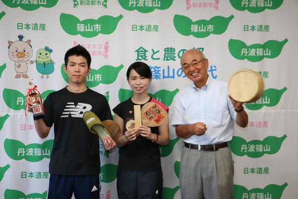 トロフィーやスリッパ・ラケットなどを手に持ったユニフォーム姿の男女ペアの選手と風呂桶を持った市長との記念写真