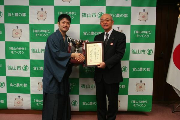 岸田 諭さんが左手にトロフィーを持ち右手で市長と握手をしており、市長が左手に額に入った賞状を持って写している写真