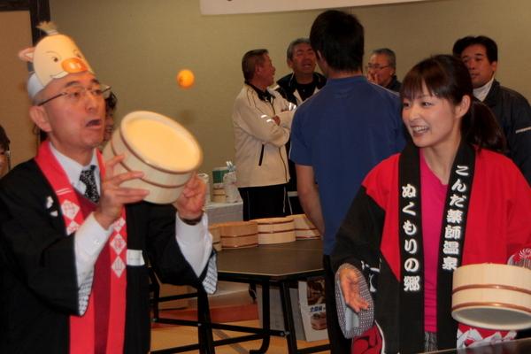 法被を着たふるさと大使の熊谷 奈美さんと法被とまるいのの紙のお面をつけた市長が桶を持ち、玉を打ち返そうとしている写真