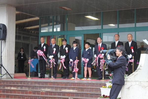 雨の中、入り口でテープカットをしている生徒と、市長、男性関係者の写真