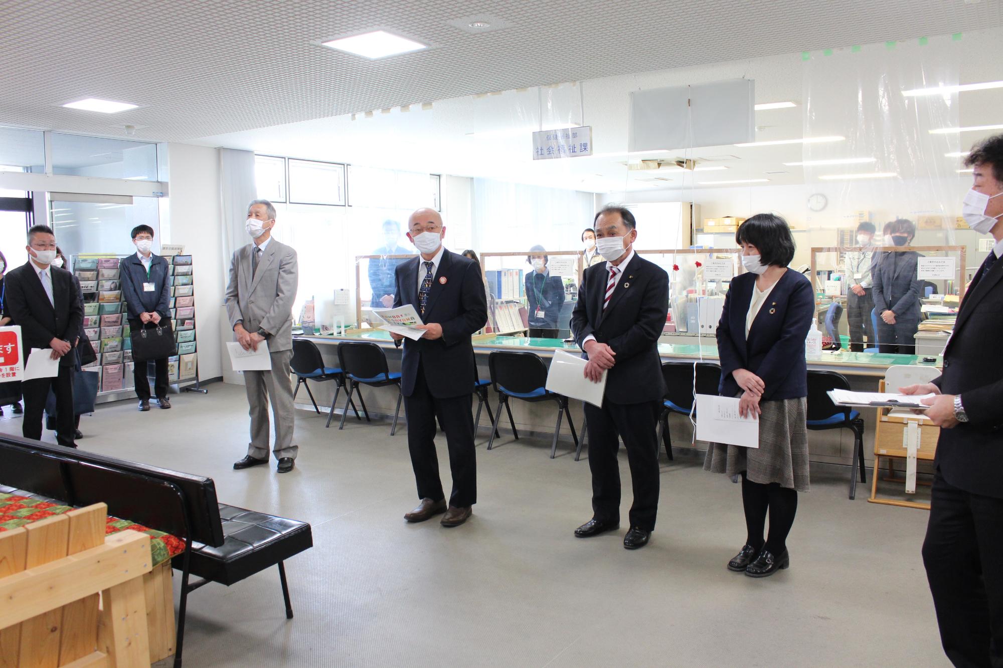 事務所フロア内で行った式典に参加された方を前に、市長が資料を手に持ってあいさつされている。