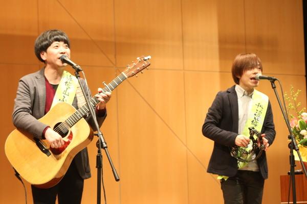 丹波篠山市のふるさと大使のタスキを肩にかけてステージ上でギターとタンバリンを手に歌っているちめいどの2人の写真