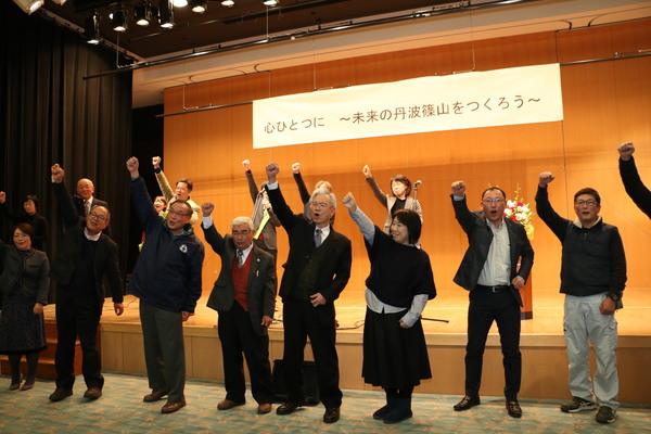 関係者たちがステージ上と下に立って右手を高く上げて叫んでいる写真