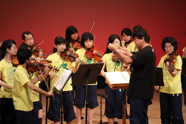 スーパーキッズ・オーケストラの学生が、佐渡さんの指揮のもとバイオリンを演奏している写真