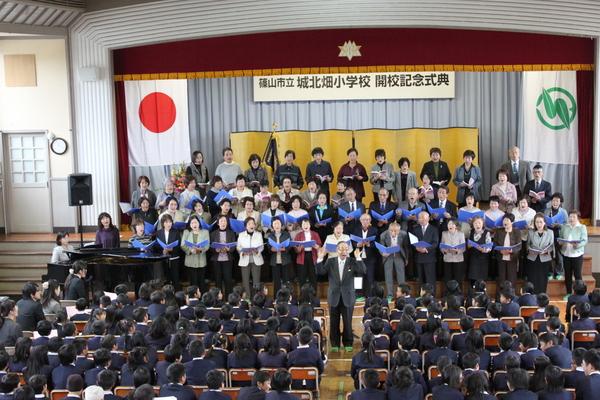 着席している生徒たちの前で、紫の紙を手に持ち、合唱している男女の写真