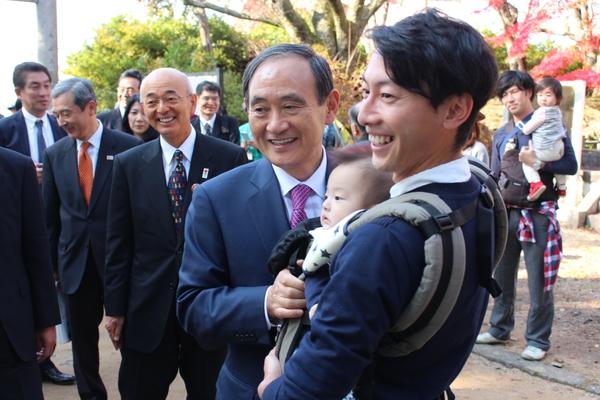 赤ちゃんを抱っこしている観光客の男性と一緒に、菅官房長官がにこやかに写真撮影に応じている様子の写真