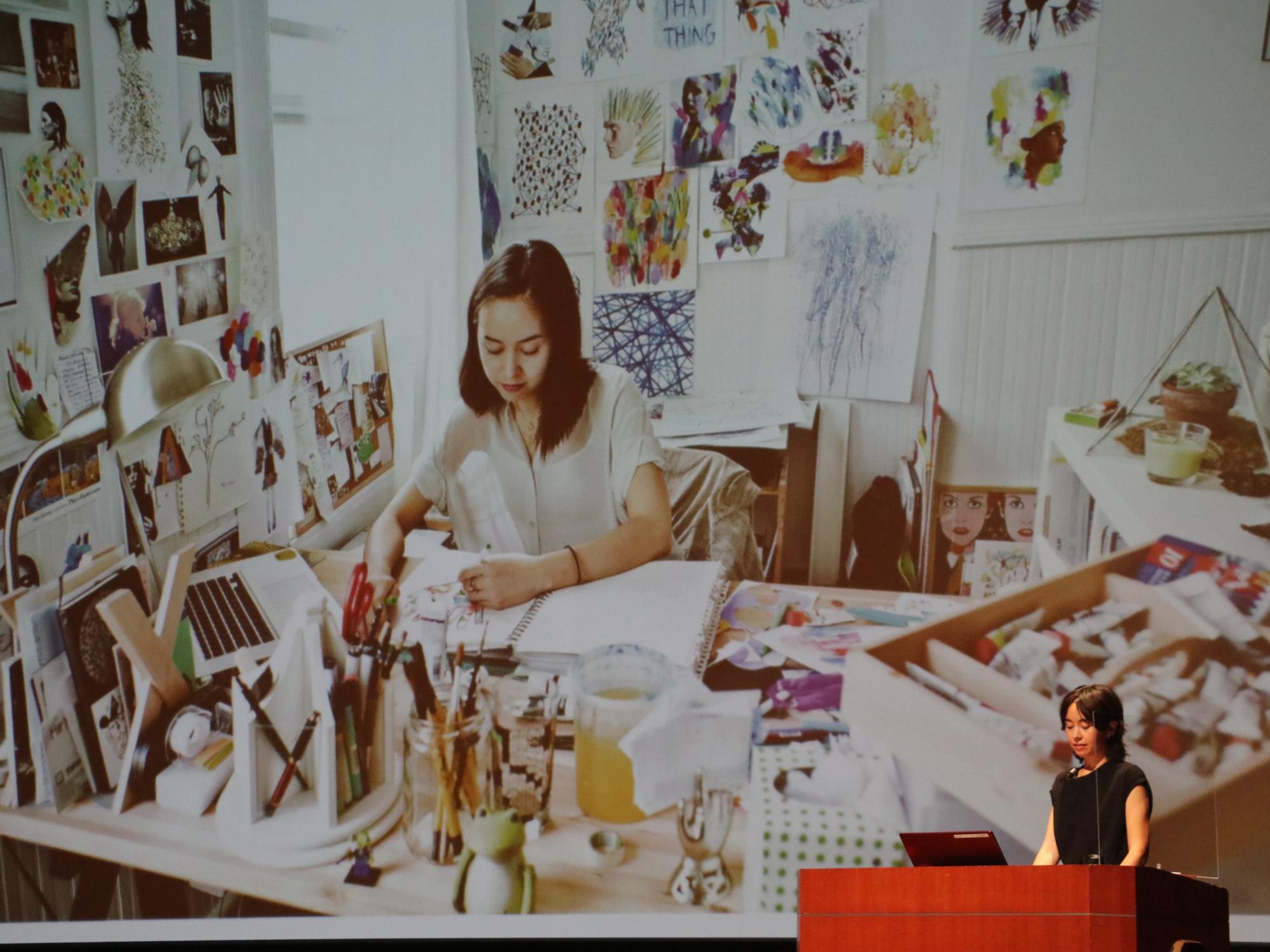 壁にデザインがたくさん貼られていて、机でデザインを考えている女性の写真をスライドに写して、女性が説明している