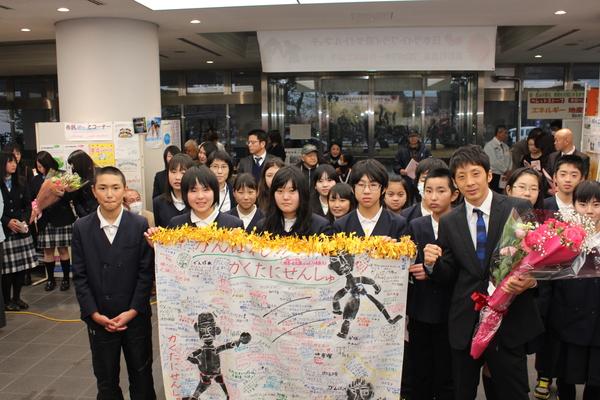 頑張れ角谷選手と書かれた、応援メッセージを子供達が持って、角谷選手は右手にガッツポーズ、左手に花束を持って記念写真