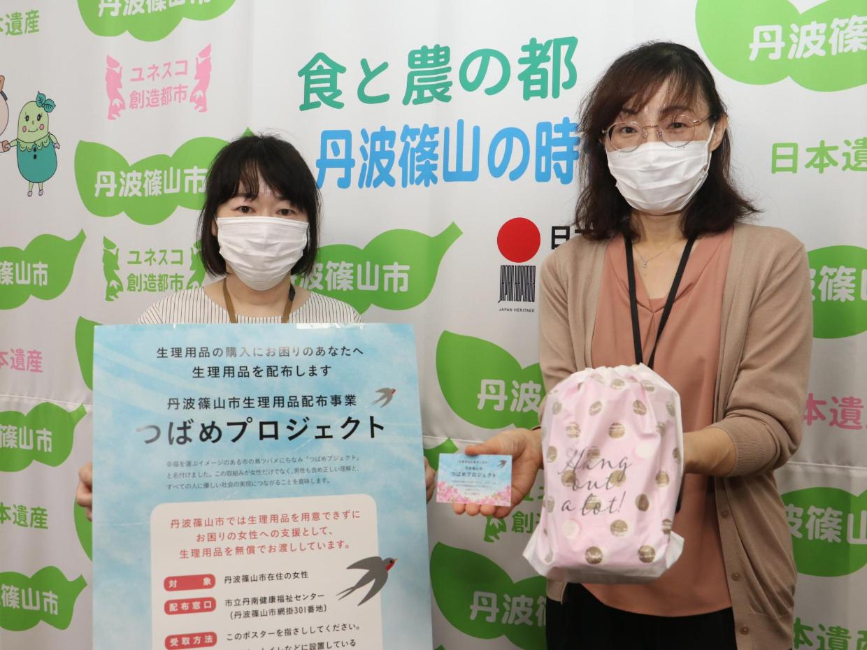 生理用品を配布するつばめプロジェクトの看板を持った女性と、水玉のポーチとつばめプロジェクトの啓発カードを手に持った女性