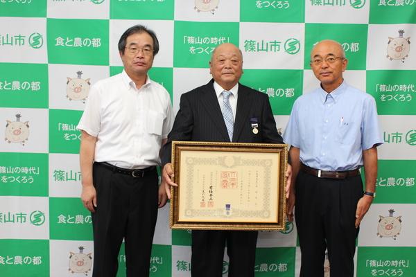 額縁に入った賞状を持った井関さんを中央に、右隣に市長、左隣に男性が立ち3人で記念撮影をしている写真