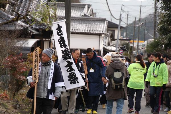 「古市義士祭」と書かれた旗を持ち歩く男性や木の札を持ち歩いている男性、ゆるキャラや黄緑色の上着を着た関係者らが道路を歩いている写真