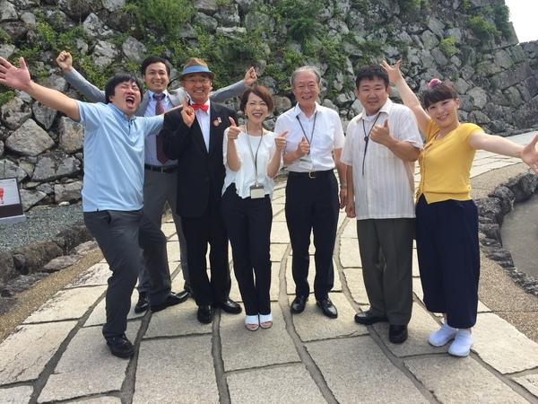 吉本新喜劇女優の森田まりこさんと帽子に蝶ネクタイ姿の市長らが石垣の前でポーズをとり記念撮影している写真