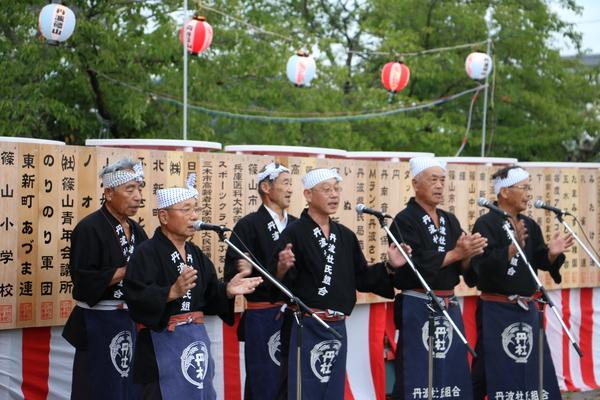 黒い法被を着て、頭にてぬぐいを巻きマイクの前に立つ男性6名の写真