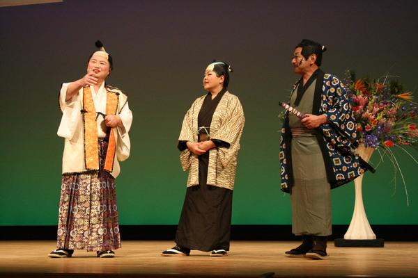 殿様の衣装を着た男性と、商人の衣装を着た男性、悪役の侍の衣装を着た男性が舞台上で寸劇をしている様子の写真