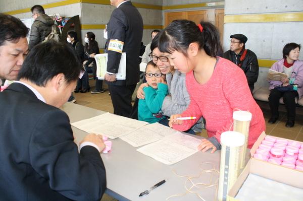 スーツの職員が説明していて、お母さんが子供を抱っこして、もう一人のお姉ちゃんが立ってペンを持っている写真