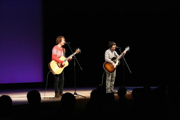 兄弟アコースティックデュオの「ちめいど」の二人がスタンドマイクを使ってギターを弾きながら歌を歌っている写真