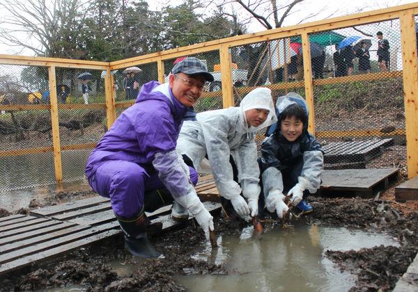 お堀の中で、カッパを着た市長と、男の子2人が種レンコンを持っている写真