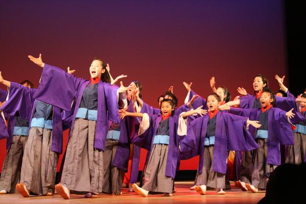 楽空間のメンバーが舞台上で紫色の羽織に茶色の袴を着て、両手を広げて歌いながら踊っている写真
