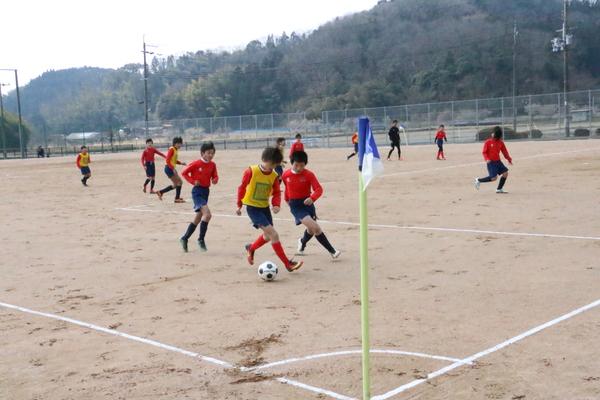 黄色いユニホームの選手が蹴っているボールを、赤いユニホームの選手が横から取ろうとしている様子の写真