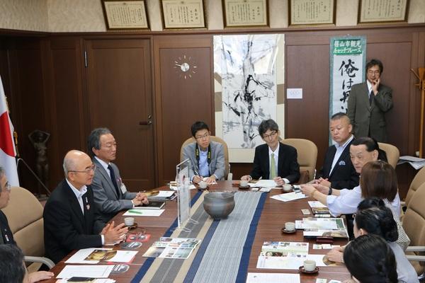 市長室の円卓の中央に壺の焼物が置かれており、市長の前に座っている水色のシャツを着た男性が話しているのを回りの人が興味深く聞いている様子の写真