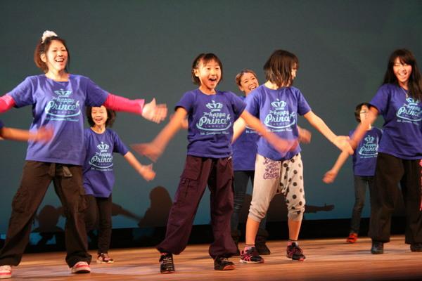 市民ミュージカルのメンバーがお揃いの紫色のティーシャツを着て、舞台上で両手を下に広げて笑顔で歌って踊っている写真