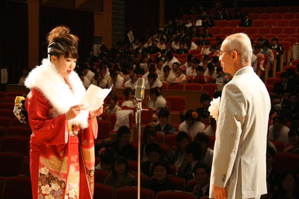 舞台上で、新成人代表 西川 早紀さんが市長の前で、誓いの言葉を述べており、客席の新成人の様子も写している写真