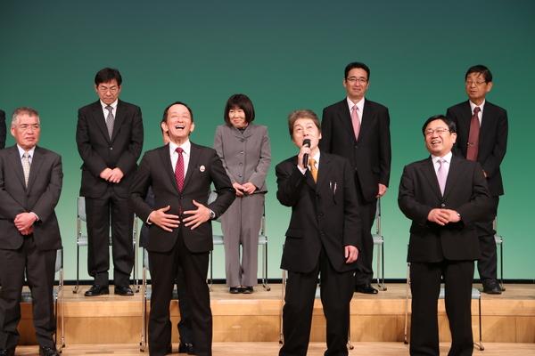 男性がマイクで歌い、横の男性が両手を腹部にあてて笑っている写真