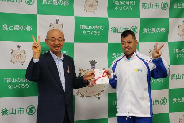 市長と岡 淳司（おか じゅんじ）さんが2人で目録をもちピースをしている写真