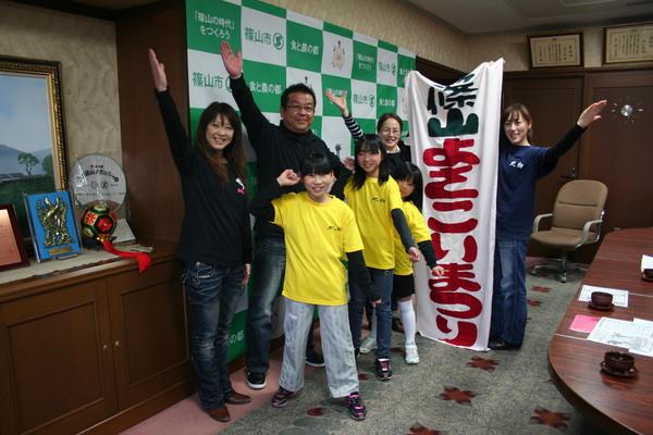 市長室でよさこいチームのメンバー5名が片手を上げてポーズをとって、後方に「篠山よさこいまつり」の垂れ幕を2名の女性が持っている写真