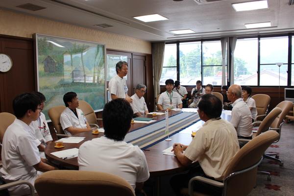 医療従事者、市長がテーブルを囲んで座り、1人の男性が立って話をしている部屋全体の様子を写した写真