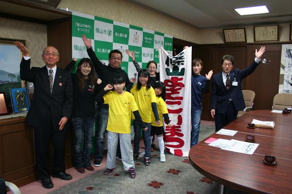 市長室でよさこいチームのメンバー5名が片手を上げてポーズをとって、後方に「篠山よさこいまつり」の垂れ幕を2名の女性が持っている左端に市長も右手を挙げてポーズをとっている写真