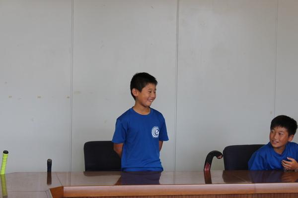 ブルーのTシャツを着た生徒が立って恥ずかしそうに挨拶をし、その横に男子生徒が座っている写真