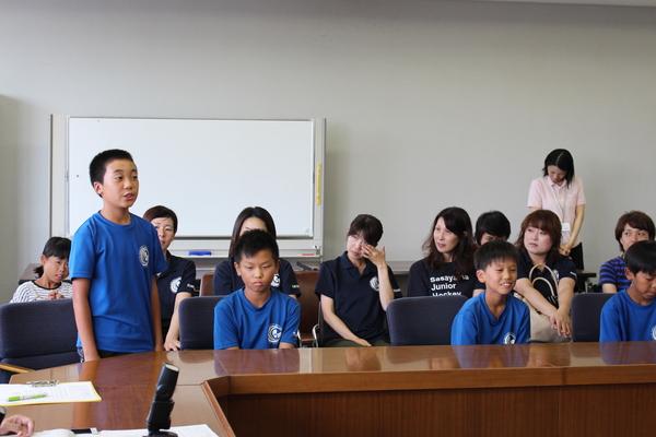 黒のTシャツを着た父兄が後ろに座っり、その前で男子生徒が立って話をしている様子を横でチームメイトが聞いている写真
