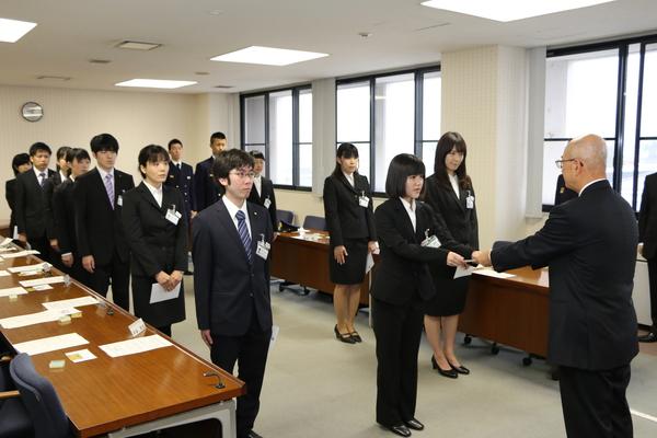 スーツ姿の新入社員が並び、市長が女性新入社員に書類を渡している写真