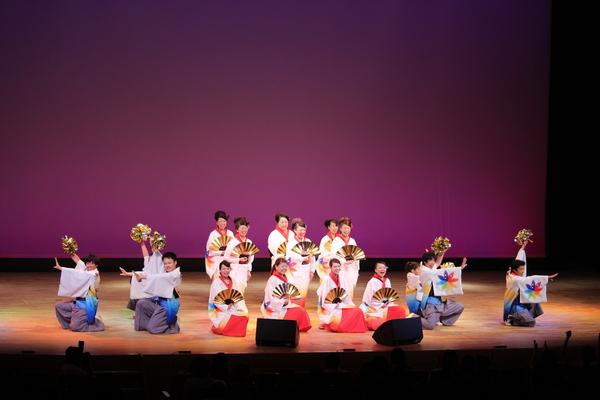 白に赤の衣装の人達は胸のあたりにゴールドのセンスをもち真ん中で前の4人がすわり後ろの6人はたちその両隣に白に紫の衣装の人達は舞台右の人は左手をまっすぐ前にあげ右手にゴールドのセンスをもち上にのばし舞台左の方々は右手をまっすぐ前にあげ左手にゴールドのセンスをもち上にのばしポーズを決めている白丹波篠山楽空間の方の写真