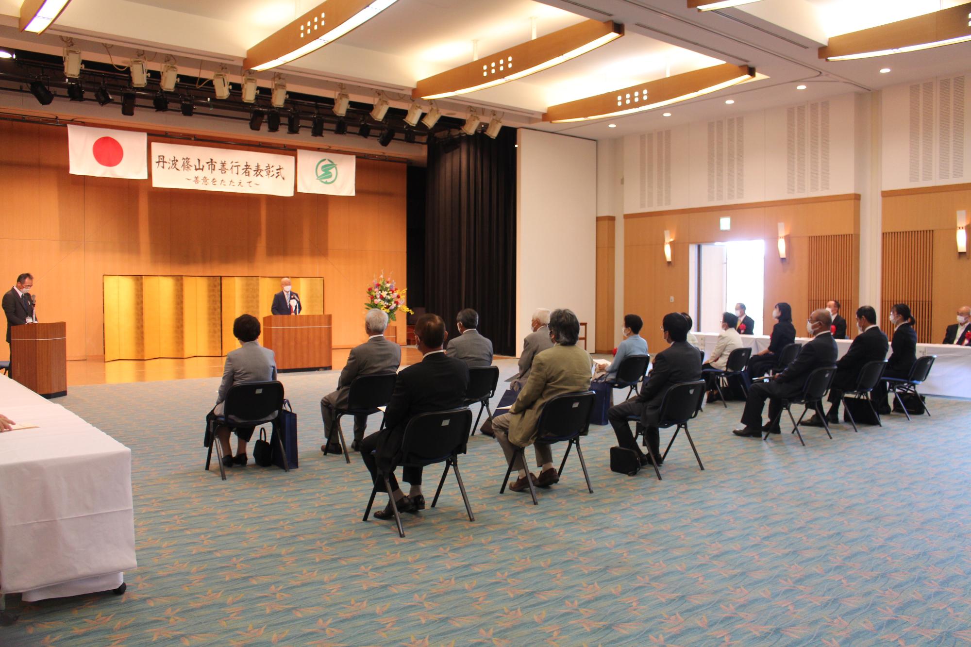会場左後方から全体を写した写真。受賞者が椅子に座り、壇上で市長が挨拶されている。