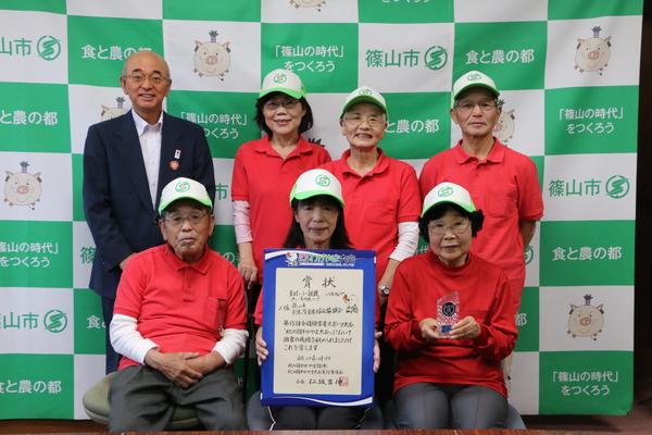 帽子にユニホーム姿の卓球バレーチームが賞状と盾を持ち、市長と記念撮影している写真