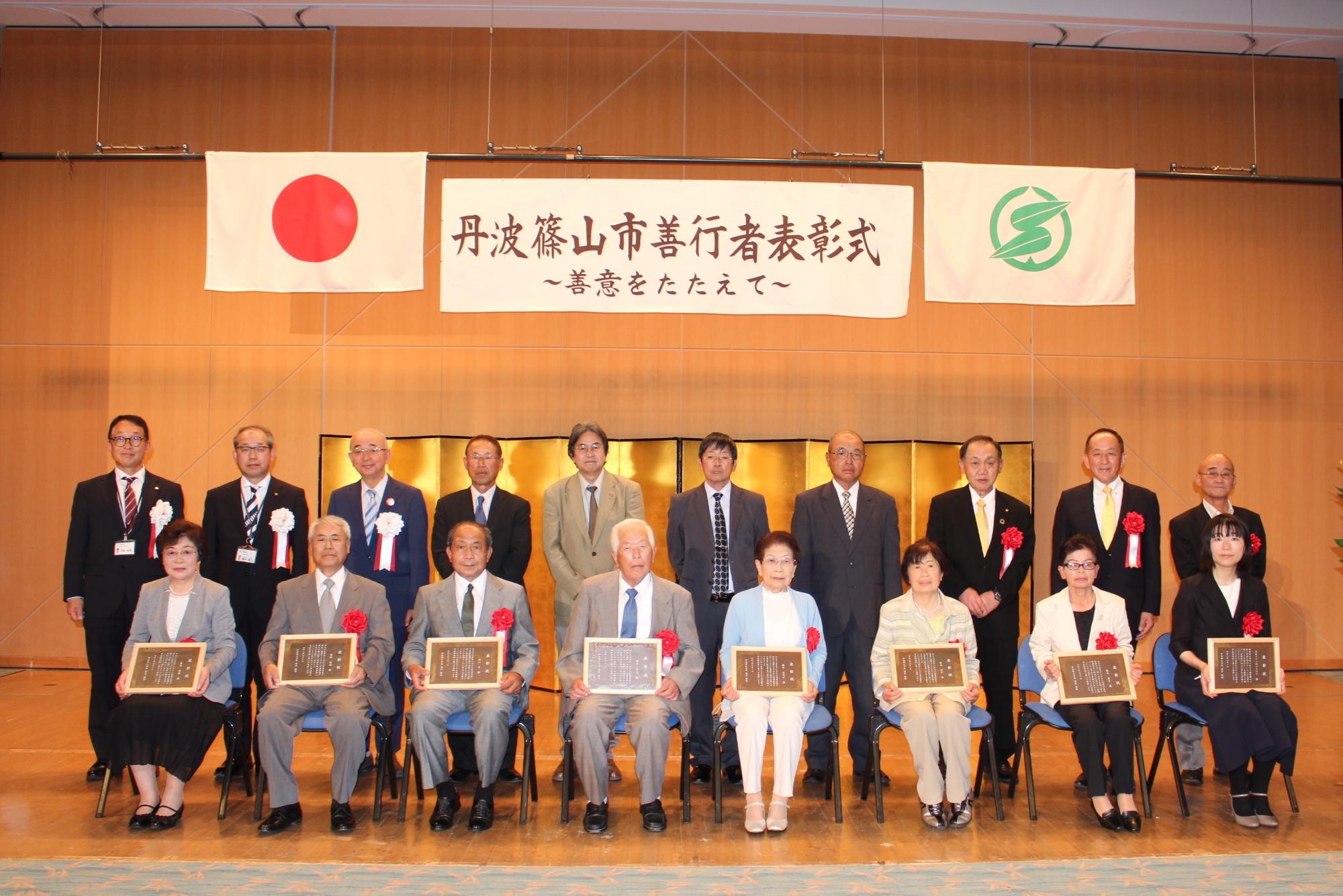 受賞者が表彰状を持って座っている。後ろに来賓が立っている。