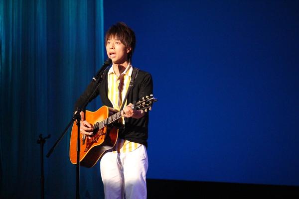 黄色と白のストライプのシャツの上に黒のジャケット、白のズボンの衣装の石田 裕之さんがギターを弾きながら、歌っている写真