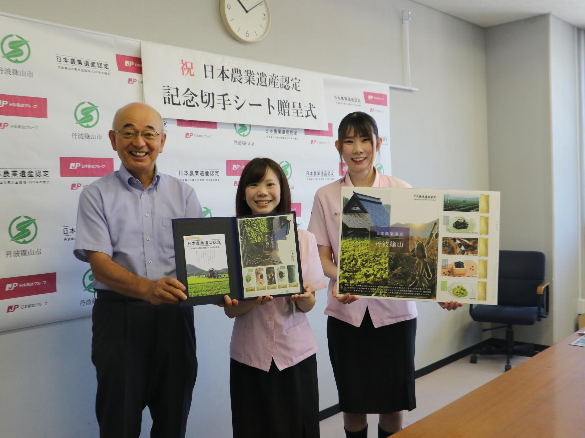 市長と女性の郵便局員が日本農業遺産記念切手シートを一緒に持ち、その隣で女性の郵便局員が日本農業遺産のパネルを持っている。