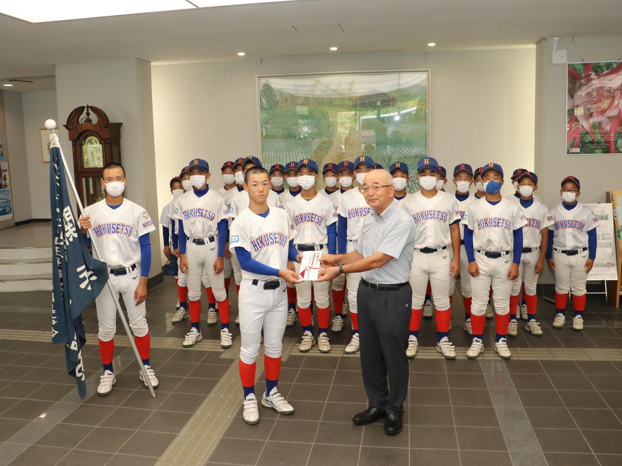 野球のユニフォームを着たシニアチームの子どもが、市長から奨励金をもらっている。後ろにチームメイトたちが立っている