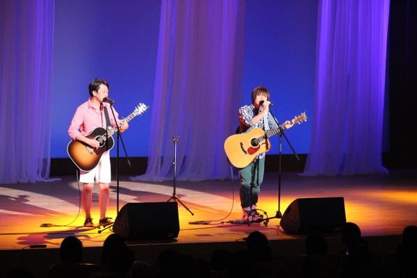 一人はピンクのシャツに白いズボン、一人は黒と白のシャツに緑のズボン姿のちめいどさん2人がギターを弾き、歌っている写真