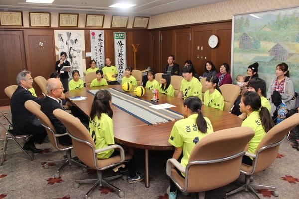 応接室に着席する黄色いユニフォームを着た女子生徒たちと市長、関係者の写真