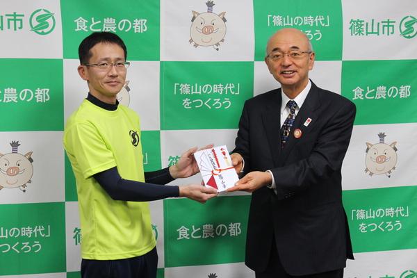 市長からのし袋を受け取る黄色いユニフォームを着た男性の写真