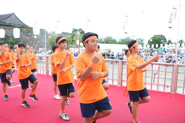 オレンジ色のシャツ、ハチマキ、靴下をはいた生徒たちが、踊っている写真